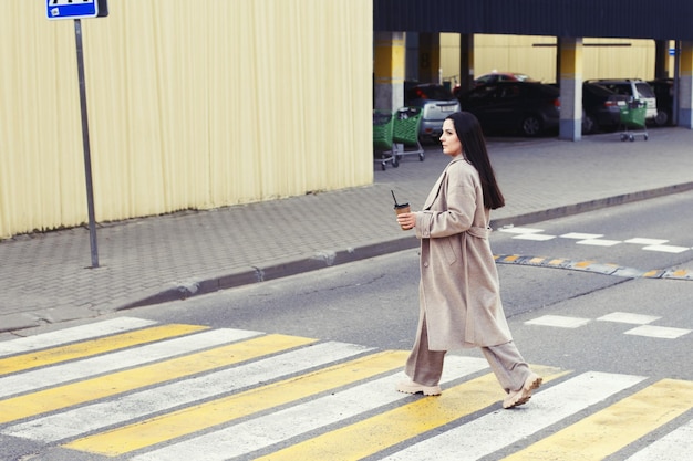 Femme tenant un café et traversant la route au passage pour piétons sur fond de ville urbaine