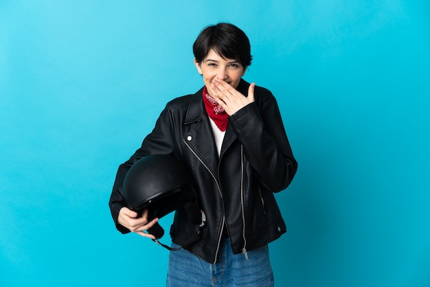 Femme tenant un casque de moto isolé sur l'espace bleu heureux et souriant couvrant la bouche avec la main