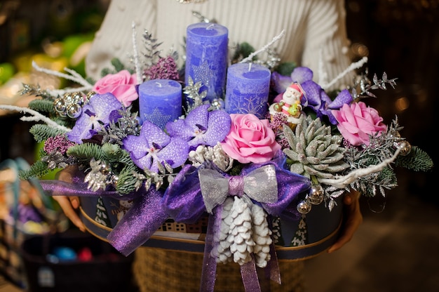 Femme tenant une composition de Noël avec des fleurs violettes et roses, des plantes succulentes, des branches de sapin et des bougies dans la boîte