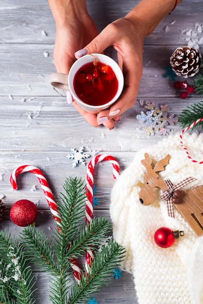 Femme tenant dans les mains du thé de Noël chaud avec canne en bonbon contre des décorations, des cerfs en bois, des cônes, de la neige, des jouets de Noël sur une planche de bois.