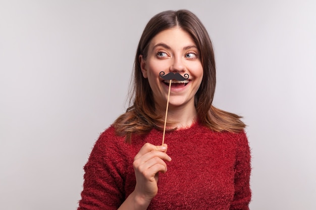 Femme tenant une fausse moustache frisée, s'amusant dans un photomaton, portant un accessoire de mascarade.