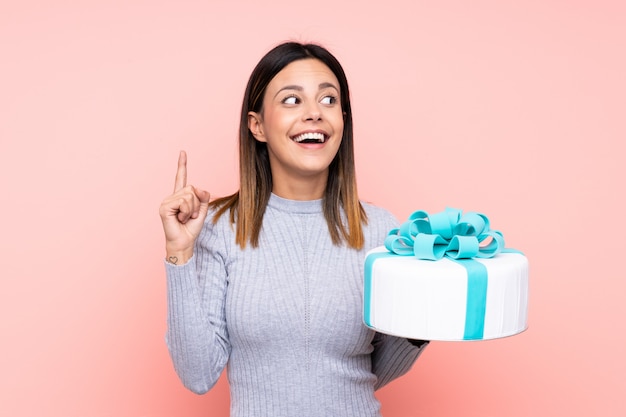 Femme tenant un gros gâteau sur un mur rose dans l'intention de réaliser la solution tout en levant un doigt