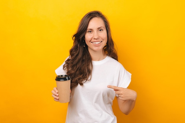 Femme tenant joyeusement une tasse de café et pendant qu'elle la pointe du doigt, elle sourit et porte une chemise blanche près d'un mur jaune