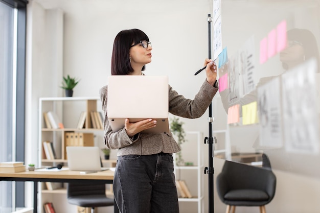 Une femme tenant un ordinateur portable et regardant un graphique financier sur un mur de verre