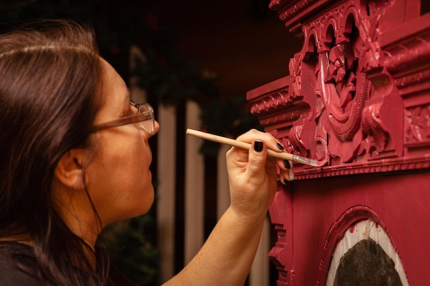 Femme tenant un petit pinceau à la main et peignant lentement une armoire en bois ornée de couleur rouge Atelier à domicile pour la rénovation de meubles Nouvelle vie pour les vieilles choses