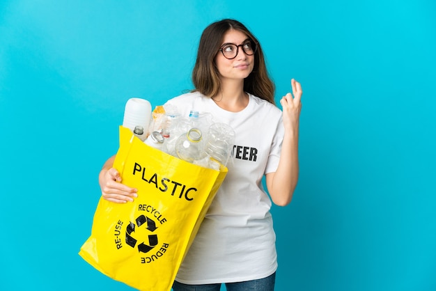 Femme tenant un sac plein de bouteilles à recycler isolé sur bleu avec les doigts qui se croisent et souhaitant le meilleur