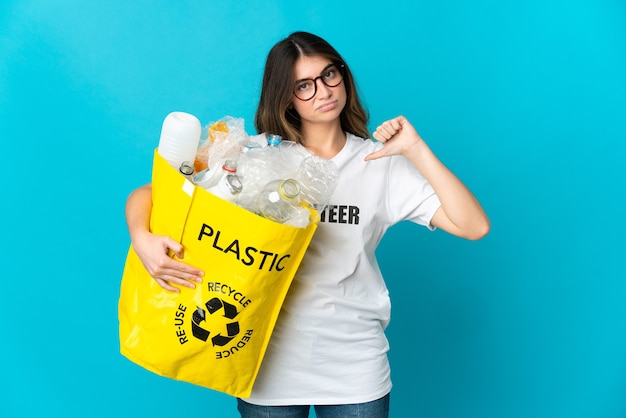 Femme tenant un sac plein de bouteilles à recycler isolé sur bleu montrant le pouce vers le bas avec une expression négative