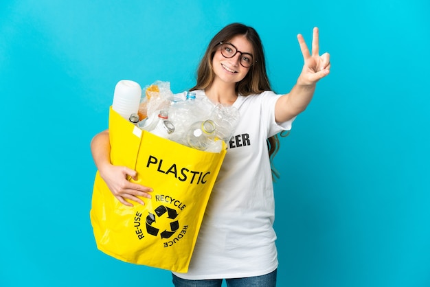 Femme tenant un sac plein de bouteilles à recycler isolé sur bleu souriant et montrant le signe de la victoire