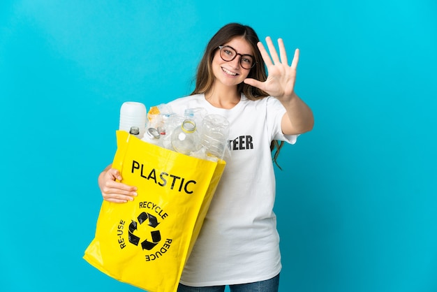 Femme tenant un sac plein de bouteilles à recycler isolé sur mur bleu en comptant cinq avec les doigts