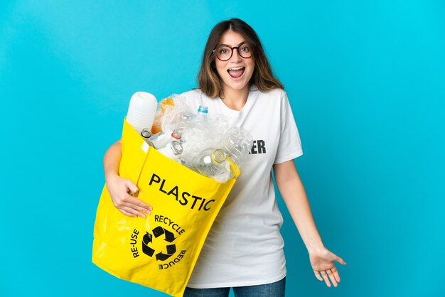 Femme tenant un sac plein de bouteilles à recycler isolé sur un mur bleu avec une expression faciale surprise