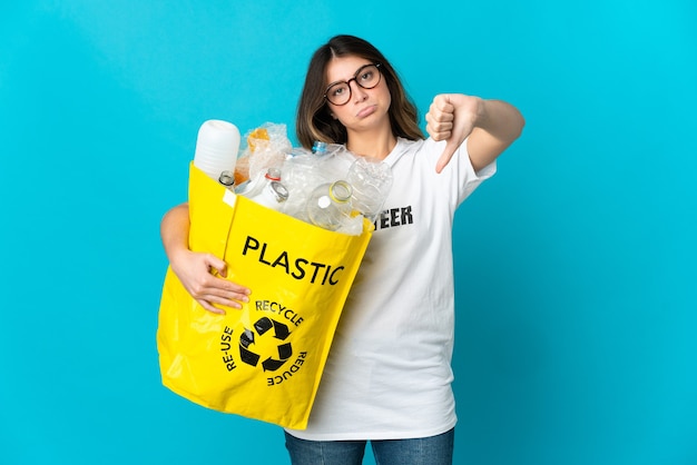 Femme tenant un sac plein de bouteilles à recycler isolé sur mur bleu montrant le pouce vers le bas avec une expression négative