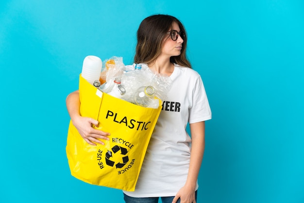 Femme tenant un sac plein de bouteilles à recycler isolé sur mur bleu à la recherche sur le côté