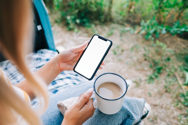 Femme Tenant Un Smartphone Avec Une Maquette D'écran Blanc, Assise Dans Une Camionnette Et Buvant Du Thé Pendant La Pause Du Voyage.