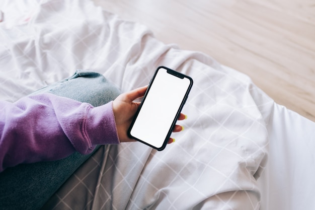 Femme tenant un smartphone avec une maquette d'écran blanc, reposant sur le lit à la maison.