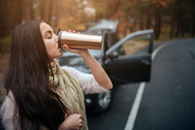 Femme tenant une tasse de café jetable à côté de la voiture. Bouchent la main. Concept d'automne. Voyage en forêt d'automne en voiture