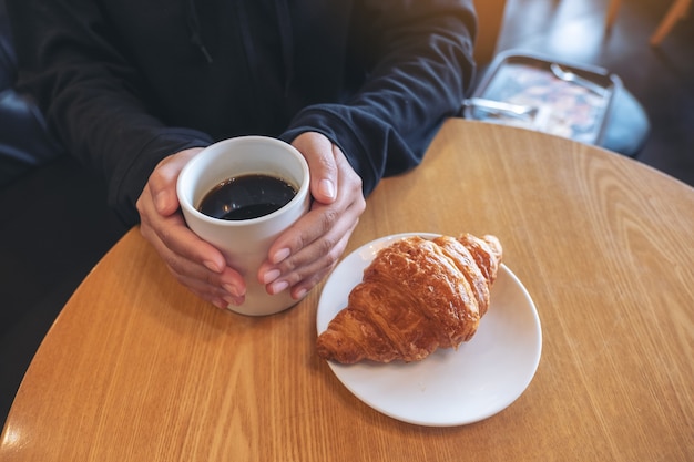 Une femme tenant une tasse de café avec un morceau de croissant dans une assiette blanche sur une table en bois au café