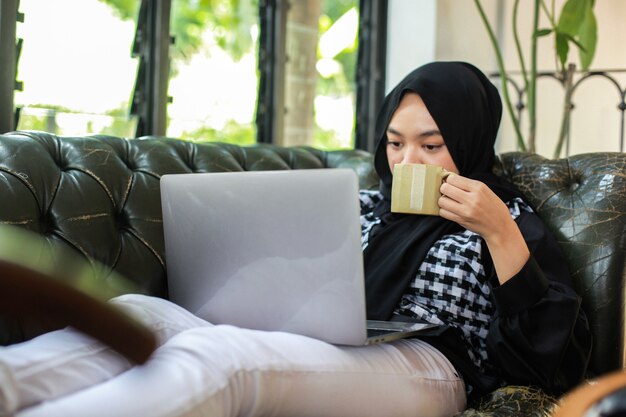 femme tenant une tasse et regardant un ordinateur portable tout en vous relaxant sur le canapé