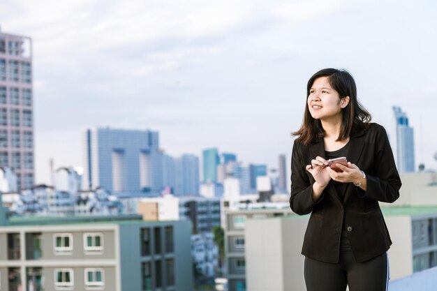 Une femme tenant un téléphone intelligent en regardant loin tout en se tenant contre le paysage urbain