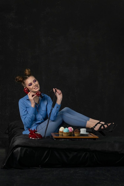 Femme tenant un téléphone rétro et des maffins Fille avec un maquillage lumineux souriant et une coiffure chignon en studio sur fond noir