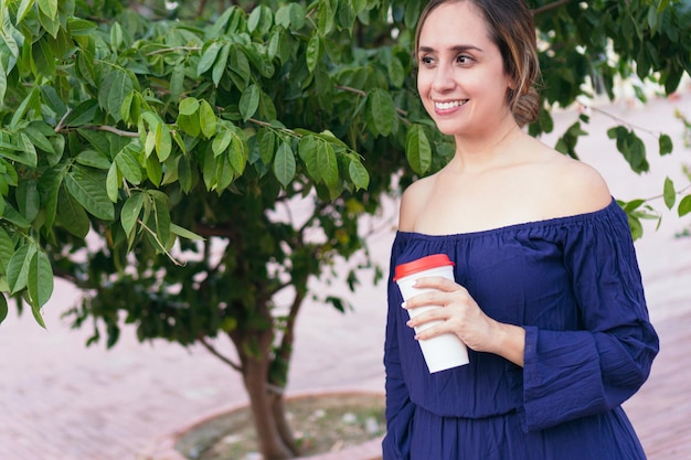 Femme tenant un verre de café et regardant sur le côté tout en se tenant dans un parc urbain