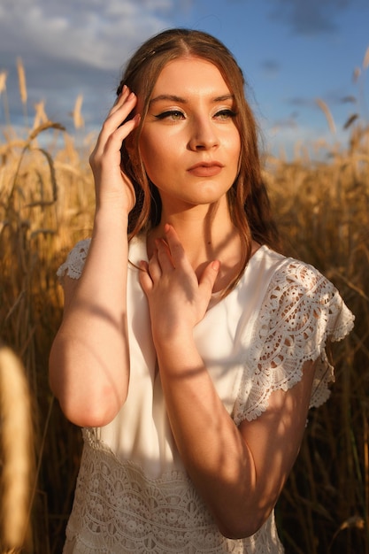 Femme tendre en robe blanche debout dans un champ de céréales avec des épillets de blé en soirée d'été