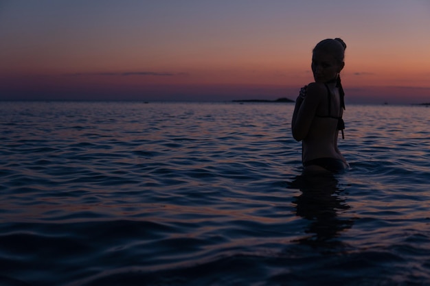 La femme tendre se tient sur la plage au coucher du soleil lumineux en mer chaude avec une silhouette