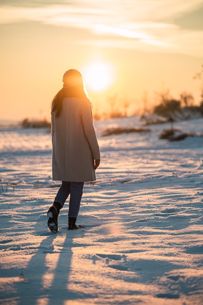 Femme en tenue chaude debout seul parmi les prairies enneigées et admirant le coucher de soleil pittoresque dans la campagne d'hiver