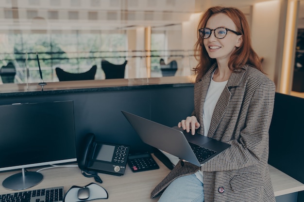 Femme en tenue décontractée assise sur un bureau dans sa cabine au bureau tout en tenant un ordinateur portable et en souriant