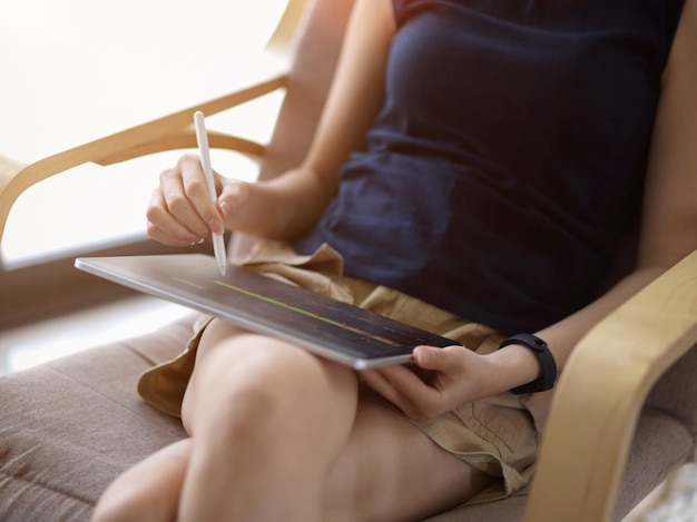 Une femme en tenue décontractée est assise dans son fauteuil confortable dans le salon de la maison et utilise une tablette