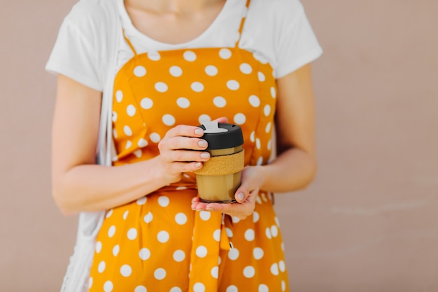 Une femme en tenue jaune tient une tasse de café réutilisable sur fond beige clair