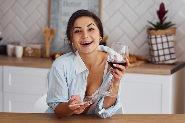 Une femme en tenue légère est dans la cuisine avec un verre de vin.