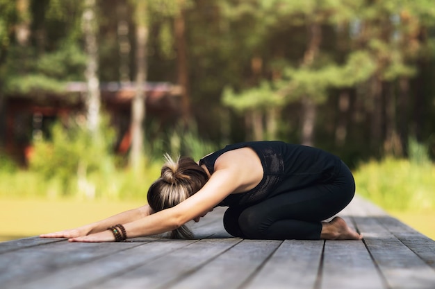 Photo femme en tenue de sport assis sur un pont en bois pratique des asanas de yoga effectuant une pose d'enfant