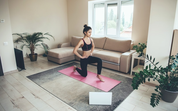 Femme en tenue de sport fait des exercices de remise en forme à la maison tout en utilisant un ordinateur portable sur un tapis rose