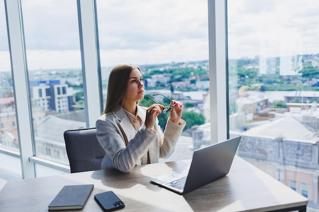 Une femme à la tête d'une entreprise à lunettes est assise devant un ordinateur portable dans son bureau avec une vue imprenable sur la ville