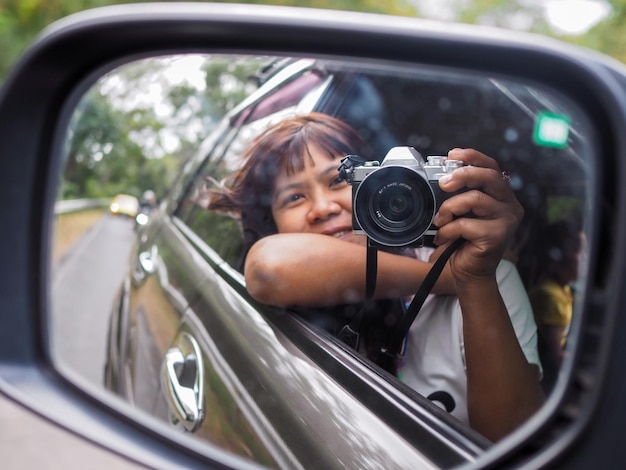 Une femme tient un appareil photo numérique et prend une photo d'elle-même souriante reflétée dans le rétroviseur de la voiture