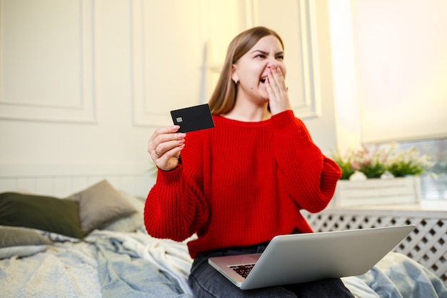 Une femme tient une carte de crédit dans ses mains, prête à faire ses courses, vêtue d'un pull rouge, assise sur le lit à la maison, une cliente de la banque montre sa carte de crédit. Achats en ligne depuis chez vous. Mise au point sélective