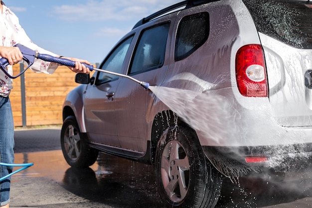 La femme tient une lance à la main et lave sa voiture à haute pression avec de l'eau et du shampoing