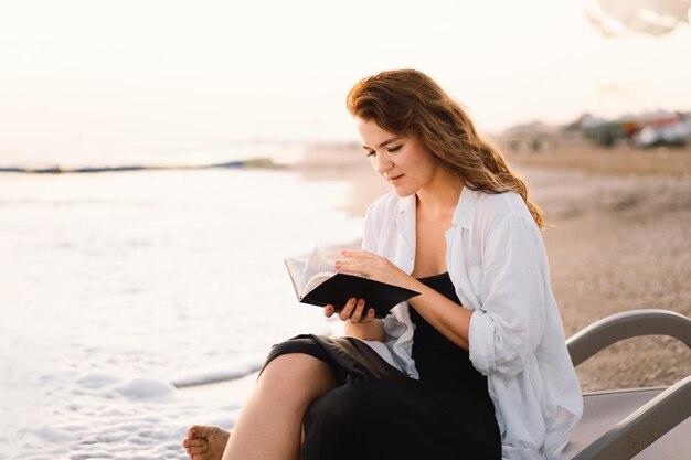 Une femme tient un livre dans ses mains en lisant le livre sur une mer au coucher du soleil