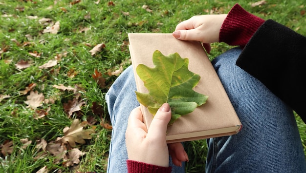 Une femme tient un livre fermé allongé sur ses genoux avec une feuille de chêne tombée en gros plan dans un parc par une chaude journée ensoleillée d'automne. Le concept de détente, de lecture et de détente seul.