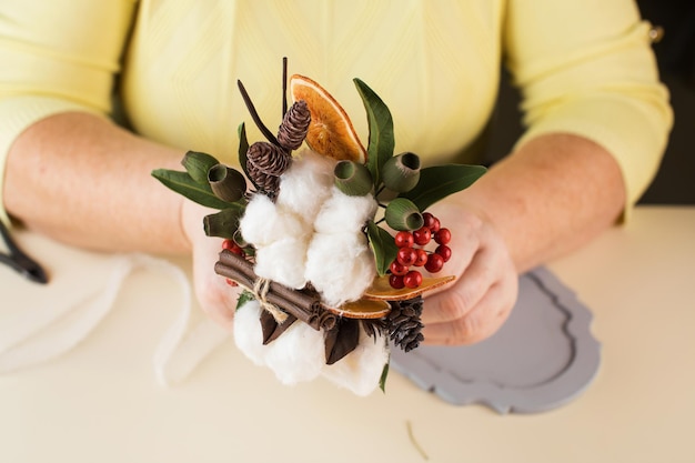 Une femme tient un petit bouquet avec des fleurs de coton et des tranches d'orange sèches au-dessus de la table