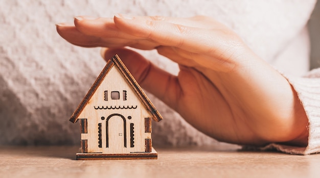 Femme tient et protège une maison en bois avec ses mains avec le soleil sur un fond rose clair. Douce maison