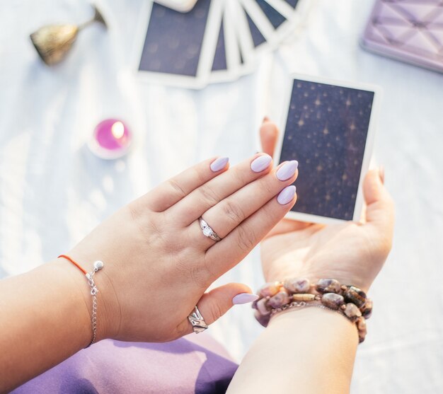 Une femme tient sa main avec des ongles violets sur une carte de tarot sur une surface blanche avec un cahier et une bougie, vue de dessus.