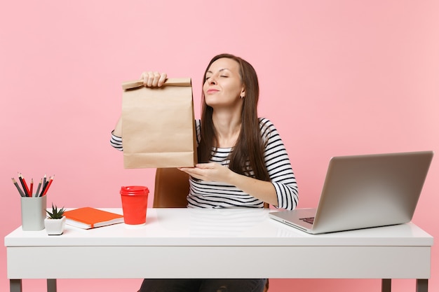 Une femme tient un sac en papier artisanal vierge et clair marron, reniflant une odeur au bureau avec un ordinateur portable isolé sur fond rose. Service de messagerie de livraison de produits alimentaires du magasin ou du restaurant au bureau.