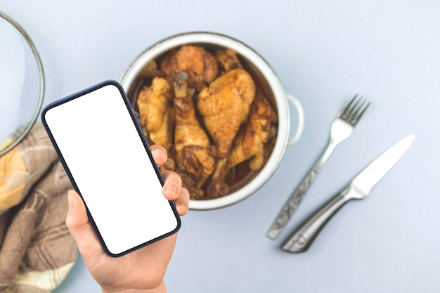 Une femme tient un téléphone portable avec un écran de maquette avec du poulet frit maison de nourriture rôtie dans une casserole, un menu de nourriture saine pour une photo de concept de restaurant, un écran blanc, une photo d'espace de copie