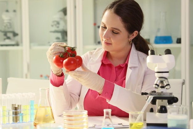 Une femme tient des tomates fraîches dans les mains assises à table près de la verrerie avec un liquide et un microscope
