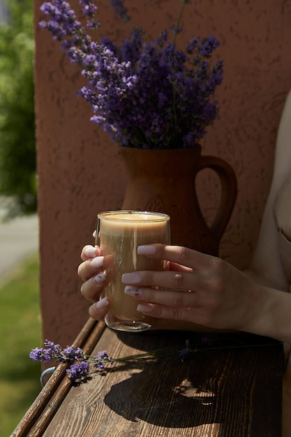 Femme tient un verre de latte sous des ombres dures Fleurs de lavande dans la cruche à l'extérieur sur la terrasse du café Pause-café esthétique