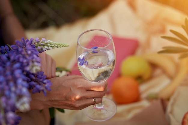 Femme tient un verre de vin avec du lupinus violet au coucher du soleil Pique-nique d'été Concept naturel de bien-être Concept de boisson ayuverdique adaptogène Consommation consciente