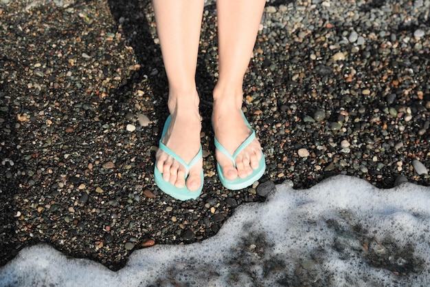 Femme en tongs élégantes sur la plage de galets près de la belle vague de mer gros plan