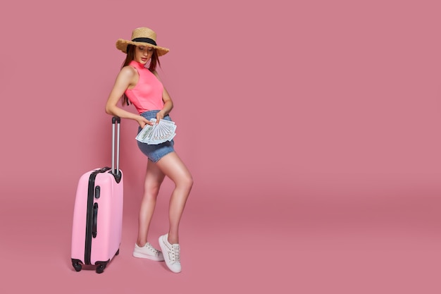 Femme de tourisme dans des vêtements décontractés d'été chapeau de paille tenant une liasse d'argent et une valise sur fond rose