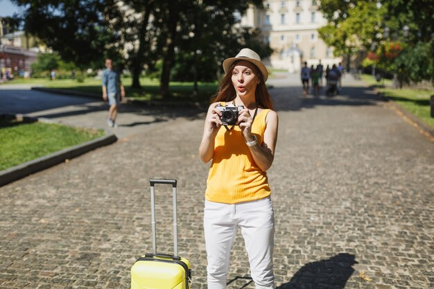 Photo femme touristique émerveillée en vêtements décontractés jaunes et chapeau avec valise prenant des photos sur un appareil photo vintage rétro en plein air. fille voyageant à l'étranger en week-end. mode de vie de voyage touristique.